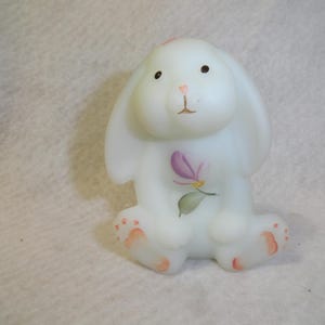 May include: A white ceramic bunny figurine with a painted pink nose, brown eyes, and pink accents on its feet. A purple and green floral design is painted on the bunny's chest. The bunny is sitting upright with its ears perked up.