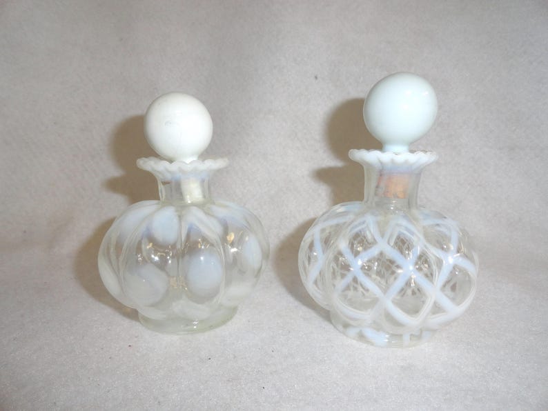 May include: Two clear glass perfume bottles with opalescent white glass accents. The left bottle features a polka dot pattern, while the right bottle has a lattice design. Both bottles have ruffled necks and round, solid white glass stoppers.