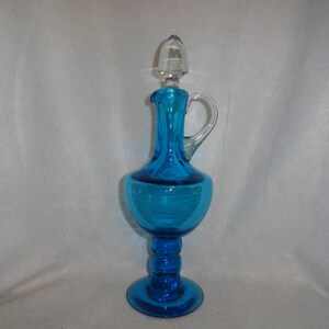 May include: A tall, blue glass bottle with a clear glass stopper and a handle. The bottle has a round body and a pedestal base.