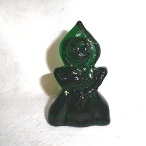 May include: A dark green glass figurine of a hooded figure, possibly a witch or a gnome. The figurine has a rounded body and a pointed hood. The details of the face are visible. The figurine is on a white surface.