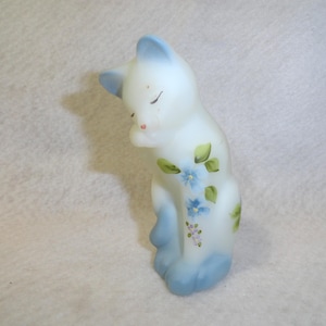 May include: A white ceramic cat figurine with blue accents on the ears, paws, and tail. The cat is decorated with hand-painted blue flowers and green leaves. The cat is in a seated position with its eyes closed.