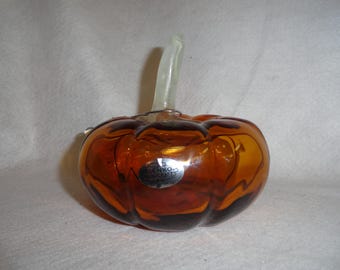 May include: A decorative glass pumpkin with a clear stem. The pumpkin is a mix of amber and brown colors with dark brown accents. A silver sticker is on the front. The pumpkin is a fall-themed decorative item.