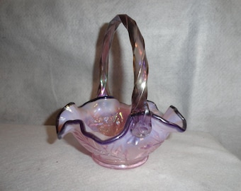 May include: A pink iridescent glass basket with a ruffled rim and a twisted handle. The basket has a floral design on the base and a purple trim. The handle is a twisted design with a metallic sheen. A decorative piece.