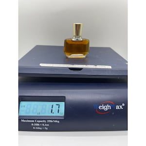 May include: A small, clear glass perfume bottle with a gold-colored cap, filled with a light amber liquid. The bottle is on a digital scale, which reads 1.7 ounces. The scale is dark blue with the brand name "Weighmax".