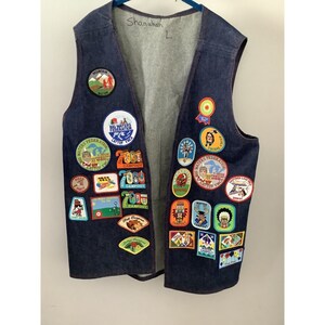 May include: A dark blue denim vest adorned with numerous colorful patches. The vest has a light gray interior and is labeled "L" and "Shanahan" in black marker. Patches feature various designs, including text and illustrations.
