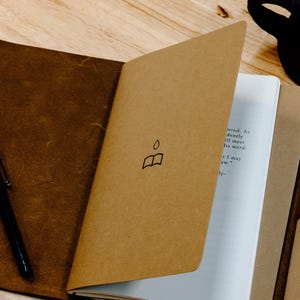 May include: A brown leather journal with a tan paper cover featuring a small black line drawing of an open book with a droplet above it. The journal is open to a white page with printed text, and a black pen rests on the dark leather cover.
