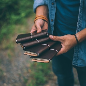 May include: A stack of four small, dark brown leather journals held together by thin white elastic bands. Each journal features a smooth, matte leather cover with visible stitching along the spine and light-colored paper pages inside.