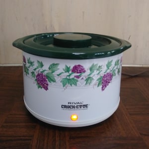 May include: A white Rival Crock-Ette slow cooker with a green lid and rim. The slow cooker has a decorative pattern of purple grapes and green leaves. The Crock-Ette logo is visible on the front, and the power indicator light is on.