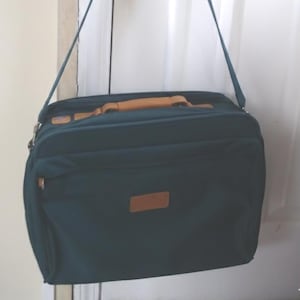May include: A dark teal briefcase-style bag with a tan handle and accents, suspended from a door handle. The bag has a rectangular shape with a front pocket and a long shoulder strap. The door is white.