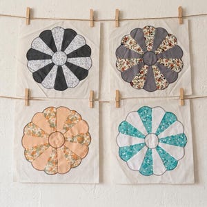 May include: Four fabric flower quilt blocks in various colors hang from a clothesline. The blocks feature petal-shaped designs in black and white, gray and floral, peach and floral, and turquoise and white. Each block is secured with a wooden clothespin.
