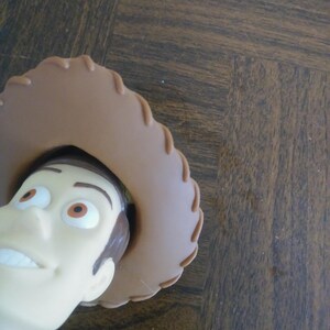 K&ouml;nnte beinhalten: Nahaufnahme des Gesichts einer Woody-Puppe, einer Figur aus dem Toy Story-Franchise. Die Puppe hat einen hellen Hautton, braune Haare und einen braunen Cowboyhut. Die Puppe tr&auml;gt ein rot-wei&szlig; gemustertes Hemd.