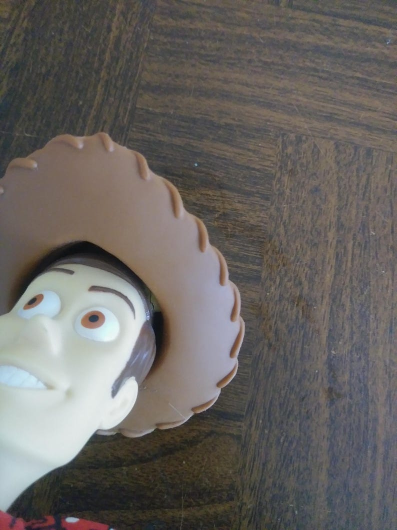 K&ouml;nnte beinhalten: Nahaufnahme des Gesichts einer Woody-Puppe, einer Figur aus dem Toy Story-Franchise. Die Puppe hat einen hellen Hautton, braune Haare und einen braunen Cowboyhut. Die Puppe tr&auml;gt ein rot-wei&szlig; gemustertes Hemd.