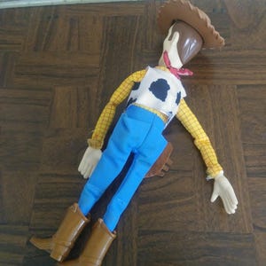 K&ouml;nnte beinhalten: Woody-Puppe, mit braunem Cowboyhut, gelbem kariertem Hemd, wei&szlig;er Weste mit schwarzen Kuhflecken, blauer Hose und braunen Stiefeln. Die Puppe hat ein rotes Halstuch und eine braune Halfter.