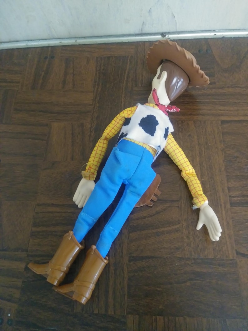 K&ouml;nnte beinhalten: Woody-Puppe, mit braunem Cowboyhut, gelbem kariertem Hemd, wei&szlig;er Weste mit schwarzen Kuhflecken, blauer Hose und braunen Stiefeln. Die Puppe hat ein rotes Halstuch und eine braune Halfter.