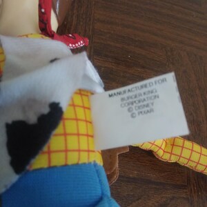 K&ouml;nnte beinhalten: Nahaufnahme einer Woody-Puppe mit gelbem kariertem Hemd, blauer Hose und einem rot-wei&szlig;em Halstuch. Das Etikett lautet "Manufactured for Burger King Corporation &copy; Disney &copy; Pixar."