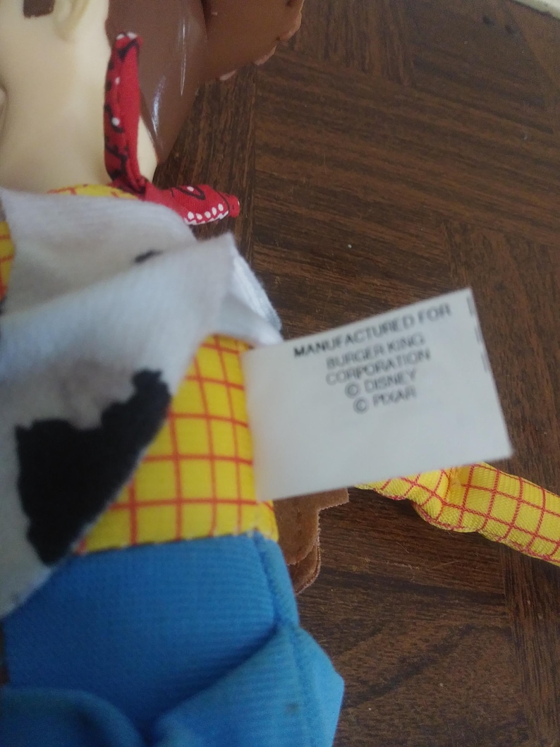 K&ouml;nnte beinhalten: Nahaufnahme einer Woody-Puppe mit gelbem kariertem Hemd, blauer Hose und einem rot-wei&szlig;em Halstuch. Das Etikett lautet "Manufactured for Burger King Corporation &copy; Disney &copy; Pixar."