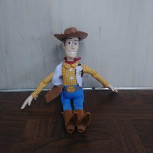 K&ouml;nnte beinhalten: Woody-Puppe aus dem Toy Story-Franchise. Die Puppe tr&auml;gt einen braunen Cowboyhut, ein gelbes kariertes Hemd, eine wei&szlig;e Weste, blaue Hosen und braune Stiefel. Die Puppe hat ein rotes Halstuch um den Hals und ein Sheriff-Abzeichen.