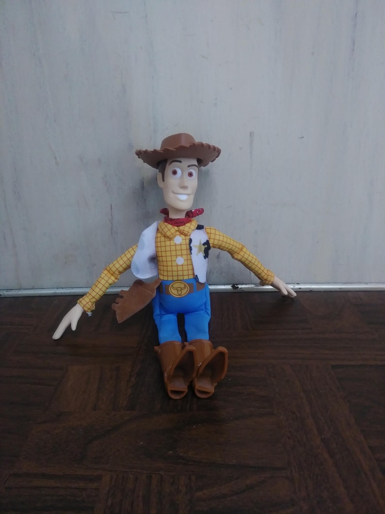 K&ouml;nnte beinhalten: Woody-Puppe aus dem Toy Story-Franchise. Die Puppe tr&auml;gt einen braunen Cowboyhut, ein gelbes kariertes Hemd, eine wei&szlig;e Weste, blaue Hosen und braune Stiefel. Die Puppe hat ein rotes Halstuch um den Hals und ein Sheriff-Abzeichen.