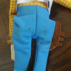 K&ouml;nnte beinhalten: Eine Woody-Puppe, von hinten gesehen, zeigt ein gelbes kariertes Hemd, eine wei&szlig;e Weste mit schwarzem Kuhfellmuster und hellblaue Jeanshosen. Die Puppe hat ein braunes Holster und eine wei&szlig;e Hand ist sichtbar.