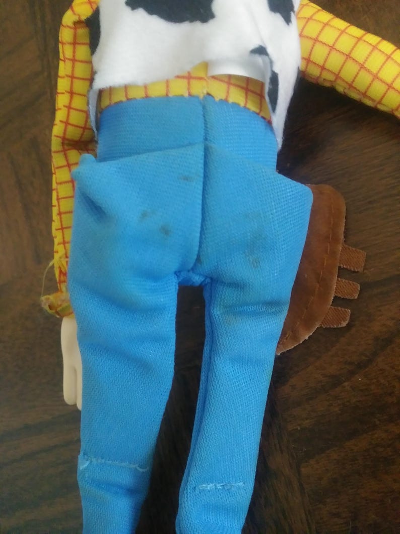 K&ouml;nnte beinhalten: Eine Woody-Puppe, von hinten gesehen, zeigt ein gelbes kariertes Hemd, eine wei&szlig;e Weste mit schwarzem Kuhfellmuster und hellblaue Jeanshosen. Die Puppe hat ein braunes Holster und eine wei&szlig;e Hand ist sichtbar.
