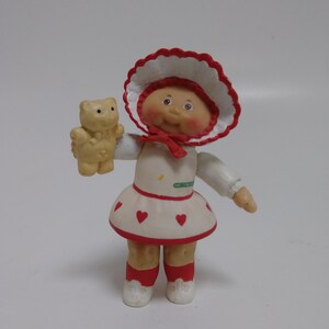 Op de afbeelding: Een vintage Cabbage Patch Kids poppenbeeldje. De pop draagt een witte jurk met rode hartaccenten en een bijpassende muts. Ze houdt een kleine, bruine teddybeer vast. De pop heeft roze wangen en een rode strik.