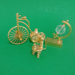 May include: A collection of miniature brass decorative items on a green background. The items include a bicycle, a clock, a small chair, and a globe with a crystal sphere. The bicycle has a large front wheel.