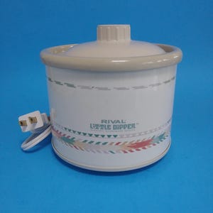 Op de afbeelding: Een witte Rival Little Dipper elektrische steengoed server met een deksel en een decoratieve band van kleurrijke driehoeken. De server heeft een beige rand en dekselhandvat. Een wit elektrisch snoer met een stekker is bevestigd.