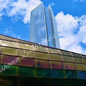 May include: A tall, blue glass skyscraper rises against a bright blue sky with white clouds. In the foreground, a curved structure features a grid of reflective, metallic, dark green and bronze-toned square panels with the text Oklahoma Backroads Photography.