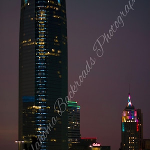 May include: A tall skyscraper with illuminated blue triangular accents at the top stands prominently in a city skyline at night. Nearby, a smaller building features a multi-colored light display on its spire, with other buildings visible.