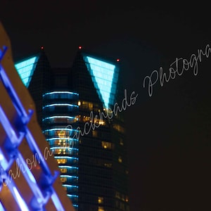May include: A night view of a skyscraper featuring a triangular illuminated top section and horizontal blue light bands, partially obscured by a foreground structure with bright blue lighting and the text Oklahoma Backroads Photography.