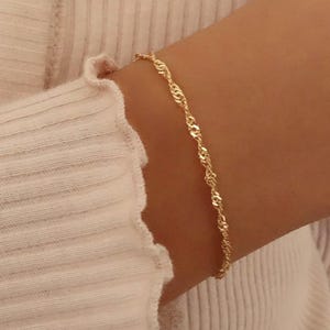 Könnte beinhalten: Ein zartes goldfarbenes Armband mit einem gedrehten Seildesign. Das Armband wird an einem Arm getragen, und der Hintergrund umfasst ein weißes geripptes Langarmshirt.