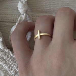Pode incluir: Um anel dourado com um design minimalista. O anel apresenta uma faixa horizontal com uma barra vertical, criando uma forma de T simples. A joia é usada em um dedo, mostrando seu estilo elegante e discreto.