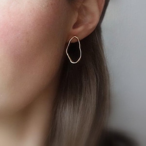 Puede incluir: Un par de pendientes de color dorado con una forma irregular y orgánica. Los pendientes son simples y modernos en diseño.