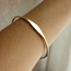 Könnte beinhalten: Ein schlichtes goldenes Armreif-Armband mit leicht gebogenem, flachem Design.