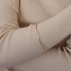 Könnte beinhalten: Ein wellenförmiges Armkettchen in Gold auf einem Arm.