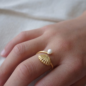 Könnte beinhalten: Ein goldfarbener Ring mit Muscheldesign und einer kleinen, runden Perle. Der Ring umschließt den Finger, wobei sich das Muschelelement am Ansatz und die Perle oben befindet. Ein zartes Schmuckstück.