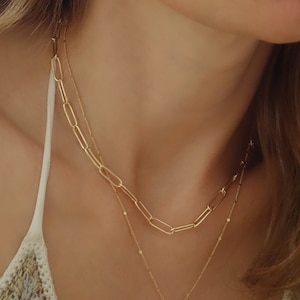 Könnte beinhalten: Eine goldfarbene Halskette mit zwei Strängen. Der obere Strang hat eine Kette aus großen, länglichen Gliedern. Der zweite Strang ist eine zarte Kette mit kleinen, gleichmäßig verteilten Perlen.
