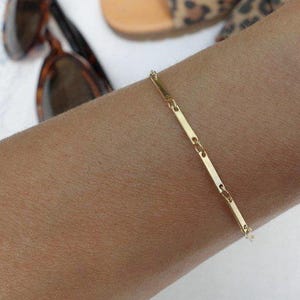 Könnte beinhalten: Ein goldenes Kettenarmband mit einem Bar-Glied-Design. Das Armband befindet sich an einem Handgelenk.