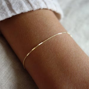Könnte beinhalten: Ein zartes goldenes Armband mit einer feinen Kette und rechteckigen Stäbchen-Akzenten. Das Armband wird an einem Handgelenk getragen, mit einem weichen, neutralfarbenen Hintergrund. Der Schmuck ist schlicht und elegant.