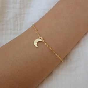 Könnte beinhalten: Ein zartes, goldfarbenes Kettenarmband mit einem kleinen, glatten Halbmond-Anhänger, der in der Mitte hängt. Das Metall hat eine polierte Oberfläche und die Kette besteht aus feinen, miteinander verbundenen runden Gliedern.
