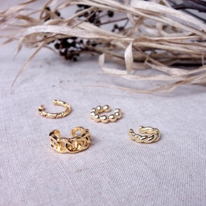 May include: Four gold ear cuffs in different styles. The ear cuffs are on a light beige fabric background.