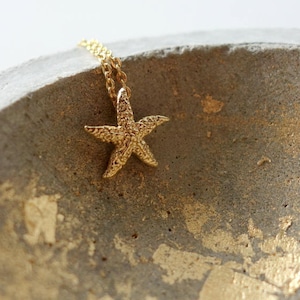May include: A gold starfish pendant necklace on a chain, resting on a textured, grey surface with gold flecks.