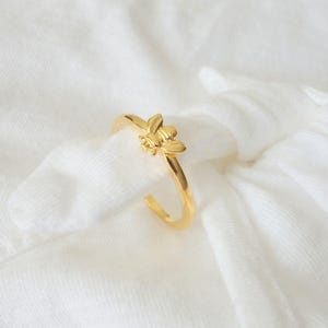 Puede incluir: Un anillo de oro con un diseño de abeja. El anillo está sobre un fondo de tela blanca.