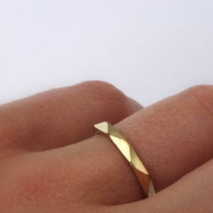 May include: A gold band ring with a geometric, faceted design. The ring is worn on a finger.