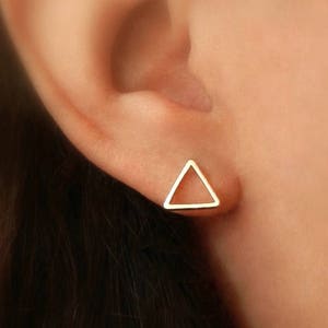 Può includere: Un orecchino a forma di triangolo in oro con un design semplice e minimalista. L'orecchino è mostrato in primo piano sull'orecchio di una persona.