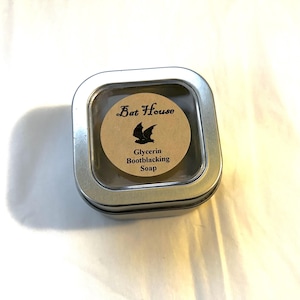 Könnte beinhalten: Eine kleine, runde, braune Seife in einer silbernen Dose mit einem durchsichtigen Deckel. Das Etikett auf der Seife lautet "Bat House Glycerin Bootblacking Soap" und zeigt eine schwarze Fledermaus-Silhouette.