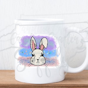 Op de afbeelding: Witte keramische mok met een kleurrijk aquarelontwerp van een konijnenkop. De achtergrond is een mix van paars, blauw en roze met glitteraccenten.