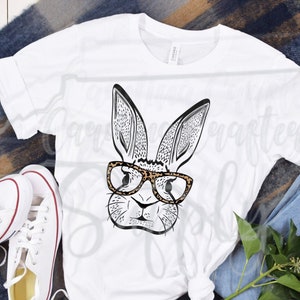 May include: White t-shirt featuring a black and white illustration of a bunny wearing leopard print glasses. The shirt is styled with white sneakers, blue jeans, and a sprig of greenery on a plaid blanket.