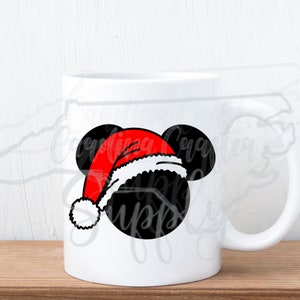 May include: White ceramic mug featuring a black Mickey Mouse head wearing a red Santa hat with a white trim and pom-pom. The mug has a curved handle and sits on a wooden surface. The design is a festive holiday theme.