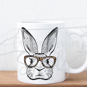 May include: White ceramic mug featuring a black and white illustration of a rabbit wearing leopard print glasses. The rabbit has long ears and a detailed face. The mug has a white handle and is set against a light background.